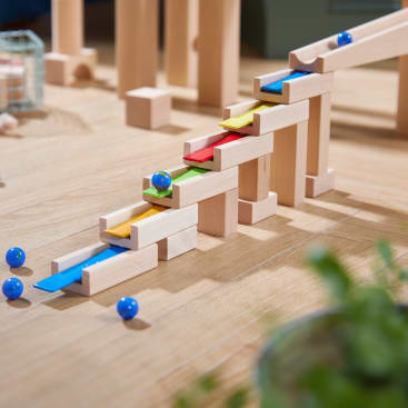 Auf einer Holzfläche ist eine hölzerne Murmelbahn mit Rampen in den Farben Blau, Gelb und Rot aufgebaut. Mehrere blaue Murmeln befinden sich auf und um die Struktur herum. Eine unscharfe grüne Pflanze erscheint in der unteren rechten Ecke.