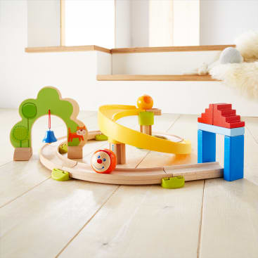 Ein Spielzeugeisenbahnset aus Holz mit einer spiralförmigen gelben Rampe, einer Kugel mit einem lächelnden Gesicht, einer blauen und roten Hausstruktur und grünen baumförmigen Bögen auf einem hellen Holzboden in einem hellen Raum.