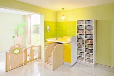 Eine hell erleuchtete, aufgeräumte Klassenzimmerecke mit einem gelben Waschbecken, einer Holztreppe in Kindergröße und einem Tor, einem hohen Regal mit durchsichtigen Behältern und grünem Wandschmuck in Form von Blättern.