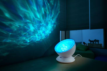 Ein kleiner weißer Projektor auf einem Tisch wirft blaue, wasserähnliche Lichtmuster auf eine dunkle Wand in einem schwach beleuchteten Raum mit geschlossenen Jalousien und einem Sofa im Hintergrund.