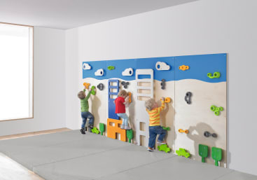Drei kleine Kinder klettern an einer bunten Indoor-Kletterwand mit verschiedenen Griffen und Formen. Der Raum hat weiße Wände, ein großes Fenster und einen gepolsterten Boden für die Sicherheit.