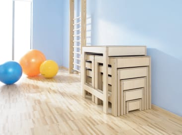 In einem Raum mit hellblauen Wänden und einer an der Wand befestigten Übungsleiter steht ein Satz verschachtelter Holzbänke auf einem hellen Holzboden neben drei bunten Gymnastikbällen.