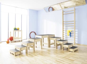Ein hell erleuchteter Raum mit hölzernen Trainingsgeräten, darunter eine Treppe, eine Wandleiter, eine Bank, ein Regal mit bunten Bällen und zwei Reifen, die an der Wand lehnen, alles auf einem hellen Holzboden.