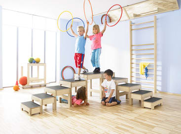Vier kleine Kinder spielen in einem hellen Turnraum mit hölzernen Klettergeräten, Stufen und bunten Hula-Hoop-Reifen. Zwei Kinder stehen oben und halten die Reifen, eines sitzt auf dem Boden, und eines liegt unter den Stufen.