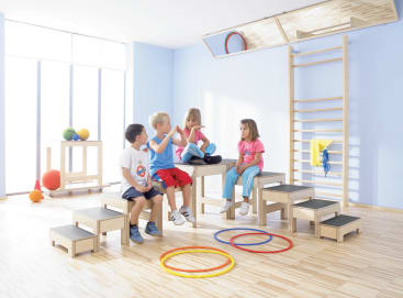 Vier kleine Kinder sitzen auf Holzstufen in einem hellen Raum mit Turngeräten, darunter bunte Reifen auf dem Boden, eine Kletterwand und Gymnastikbälle auf einem Wagen in der Nähe großer Fenster.