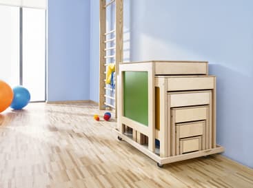 Ein hölzerner Aufbewahrungswagen mit Schubladen und einer grünen Seitenwand steht auf einem hellen Holzboden in einem Raum mit blauen Wänden, Fitnessgeräten, bunten Bällen und hellem Tageslicht.