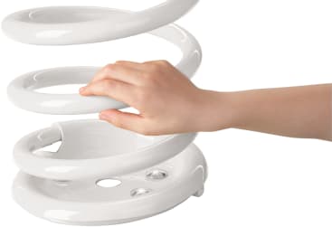 Eine Kinderhand, die durch die Öffnung eines weißen, spiralförmigen Objekts mit glatter, glänzender Oberfläche greift, vor einem unifarbenen Hintergrund.