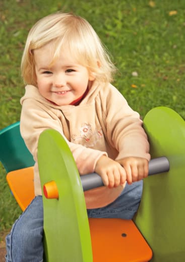 Ein kleines Kind mit blondem Haar, das einen beigen Kapuzenpulli und blaue Jeans trägt, sitzt auf einer grün-orangefarbenen Spielplatzwippe und hält sich an einer Metallstange fest, während im Hintergrund Gras zu sehen ist.