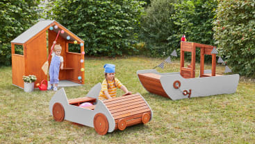 Zwei Kinder spielen im Freien auf einer Wiese mit hölzernen Spielgeräten in Form eines Hauses, eines Autos und eines Bootes. Die Strukturen sind braun und grau, und es gibt Bäume und Sträucher im Hintergrund.