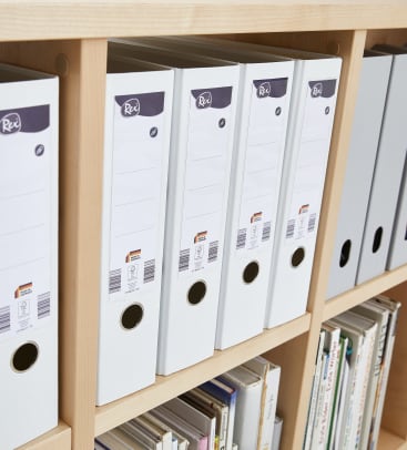 Eine Reihe weißer Aktenordner mit Blanko-Etiketten ist ordentlich auf einem Holzregal angeordnet, weitere Ordner und Bücher sind in den Regalen darunter gelagert.
