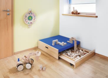 Ein aufgeräumtes Kinderspielzimmer mit hellem Holzboden. In der Ecke steht eine Holzkiste mit einem blauen Kissen; die Schublade ist geöffnet und mit Holzklötzen gefüllt. Holzspielzeug und ein dekoratives Wandobjekt sind ebenfalls zu sehen.