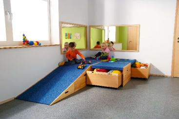 Drei kleine Kinder spielen mit Spielzeug auf einer Rampe und einem Podest mit blauem Teppichboden in einem Raum mit hellen Wänden, Spiegeln und hölzernen Spielzeugschränken. Auf dem Podest befinden sich bunte Bälle und Klötze.
