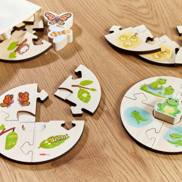 Vier runde Holzpuzzles mit Abbildungen von Tieren und Insekten in verschiedenen Lebensstadien sind auf einem Holzboden angeordnet. Die Teile zeigen Frösche, Schmetterlinge, Raupen, Eier und Blätter. Einige Puzzles sind unvollständig.