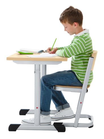 Ein Junge in grün-weiß gestreiftem Hemd und Jeans sitzt an einem Holzschreibtisch und schreibt mit einem grünen Bleistift in ein Notizbuch. Er ist auf seine Arbeit konzentriert und sitzt auf einem einfachen Stuhl.
