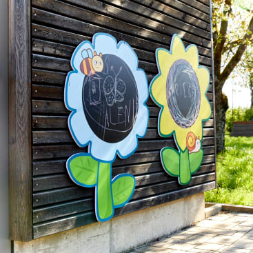 Zwei blumenförmige Kreidetafeln mit Cartoon-Bienen- und Schneckendekoration sind an einer Holzwand im Freien angebracht. Jede Tafel ist mit Kreidezeichnungen und Schrift von Kindern versehen. Im Hintergrund sind Sonnenlicht und Grünpflanzen zu sehen.