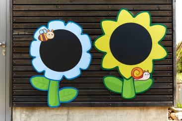 Zwei große blumenförmige Tafeln sind an einer Holzwand angebracht. Eine Tafel ist blau und weiß mit einer Cartoon-Biene, die andere gelb mit einer Cartoon-Schnecke; beide haben große schwarze Zentren und grüne Blätter am unteren Rand.