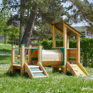 Ein hölzernes Spielgerät auf einer Wiese mit einer kleinen Rutsche, einer Treppe, Kletterstangen und einer überdachten Plattform, umgeben von Bäumen und Grünpflanzen an einem sonnigen Tag.