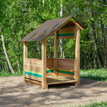 Ein kleines hölzernes Spielhaus mit einem schrägen Dach und offenen Wänden steht auf sandigem Boden. Die Struktur weist grüne und blaue horizontale Latten auf. Im Hintergrund sind üppige grüne Bäume und Gras zu sehen.
