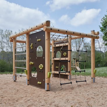 Ein hölzernes Spielplatz-Klettergerüst mit Leitern, Seilen und Kletterwänden mit Haltegriffen steht auf sandigem Boden in einem Park mit Gras und Bäumen im Hintergrund.