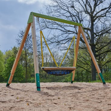Eine Schaukel mit einem runden Korbsitz und gelben Seilen steht auf einem sandigen Spielplatz, umgeben von Bäumen und Grünpflanzen unter einem teilweise bewölkten Himmel.