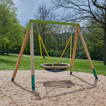 Eine runde Nestschaukel mit einem geflochtenen Sitz ist an einem Holzgestell in einem sandigen Spielbereich aufgehängt, der an einem sonnigen Tag von Gras und Bäumen umgeben ist.