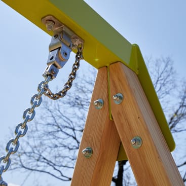Nahaufnahme einer Metallkette und einer Halterung, die an einem hölzernen A-Rahmen befestigt sind, wahrscheinlich Teil einer Schaukel, mit Schrauben zur Sicherung des Rahmens und einem klaren Himmel im Hintergrund.
