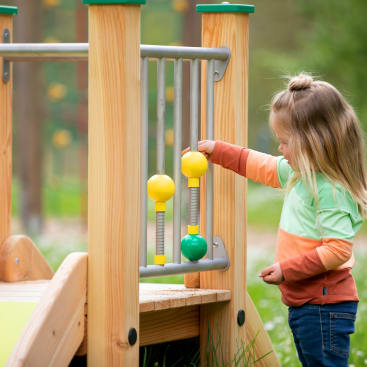 Ein kleines Kind mit langen blonden Haaren und einem bunten langärmeligen Hemd steht auf einem Spielplatz aus Holz und spielt mit gelben und grünen Drehknöpfen, die an Metallstangen befestigt sind.