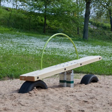 Eine hölzerne Wippe mit einem grünen Metallgriff steht auf einem sandigen Spielplatz, der von Reifen gestützt wird, mit Gras, kleinen weißen Blumen und Bäumen im Hintergrund.