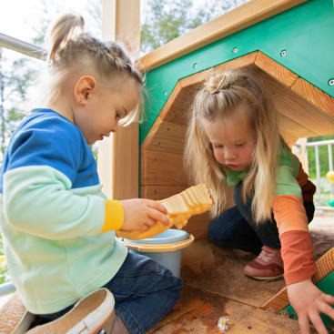 Zwei kleine Kinder mit hellem Haar spielen mit Sand und Spielzeug in einem hölzernen Spielhaus im Freien. Ein Kind benutzt eine Spielzeugbürste, während das andere sich nach vorne lehnt, beide konzentriert auf ihre Tätigkeit.