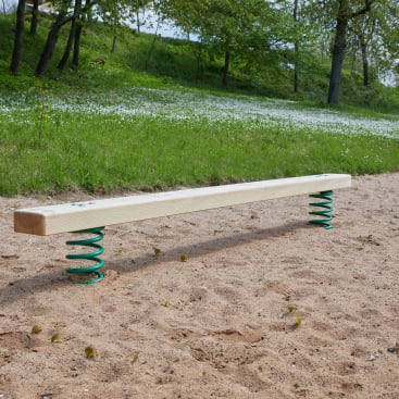 Ein hölzerner Schwebebalken mit grünen Metallfedern an beiden Enden ist auf einer sandigen Fläche in einem Park im Freien installiert, mit Gras und Bäumen im Hintergrund.
