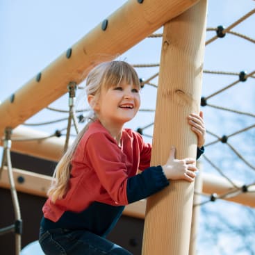Ein junges Mädchen mit blonden Haaren klettert lächelnd auf eine Holzstange auf einem Spielplatz im Freien. Sie trägt ein rot-marinefarbenes langärmeliges Hemd und Jeans. Die Spielplatzstruktur umfasst Seile und Holzbalken.