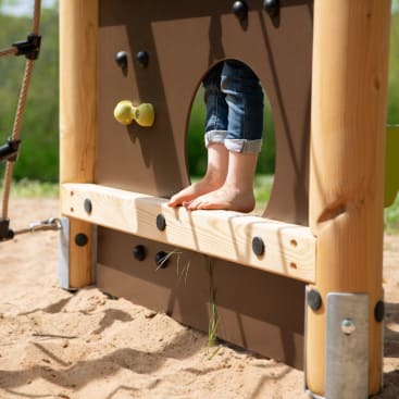 Ein Kind mit hochgekrempelten Jeans steht barfuß auf einem hölzernen Spielplatz, so dass nur seine Beine und Füße zu sehen sind. Der Boden darunter ist sandig mit etwas Gras.