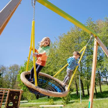 Zwei Kinder spielen an einem sonnigen Tag auf einer großen runden Schaukel in einem Park. Ein Kind sitzt, während das andere steht, umgeben von grünen Bäumen und Gras unter einem klaren blauen Himmel.