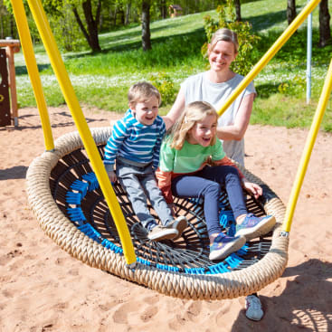 Ein Erwachsener schiebt zwei lächelnde Kinder, die zusammen auf einer großen runden Schaukel auf einem Spielplatz sitzen. Die Schaukel hat gelbe Seile und der Boden ist mit Sand bedeckt. Im Hintergrund sind Bäume und Gras zu sehen.