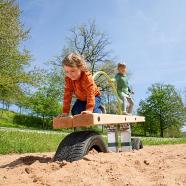 Zwei Kinder spielen auf einer hölzernen Spielplatzwippe mit Reifen darunter, die in einem sandigen Bereich steht. Ein Kind krabbelt auf der Wippe, während das andere dahinter steht, an einem sonnigen Tag im Freien mit Bäumen und Gras im Hintergrund.