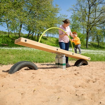 Ein Erwachsener und ein Kind stehen auf einem sandigen Spielplatz neben einer hölzernen Wippe mit Reifen darunter. Der Erwachsene hält die Hand des Kindes, während das Kind auf der Wippe steht und einen grünen Schlauch hält. Im Hintergrund sind Bäume und Gras zu sehen.