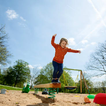 Ein kleines Kind in einem orangefarbenen gestreiften Hemd und Jeans balanciert auf einer federnden Wippe auf einem Spielplatz, während im Hintergrund bei klarem Himmel Spielzeug und Schaukeln zu sehen sind.