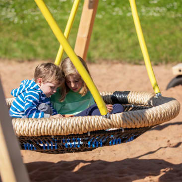 Zwei kleine Kinder sitzen zusammen auf einer großen runden Seilschaukel auf einem Spielplatz. Die Schaukel hängt an gelben Seilen über einer sandigen Fläche, und beide Kinder schauen nach unten, möglicherweise auf etwas in ihrem Schoß.