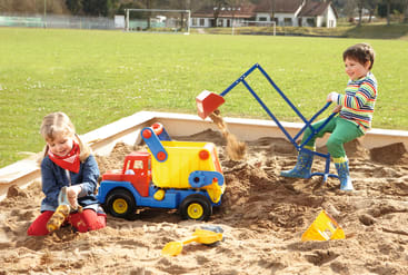 Zwei Kinder spielen in einem Sandkasten. Eines gräbt mit einer Spielzeugbagger-Maschine, das andere sitzt und hält einen Stock. Im Sand befinden sich ein großer Spielzeug-Kipplaster und andere kleine Sandspielzeuge. Im Hintergrund ist eine grasbewachsene Wiese zu sehen.