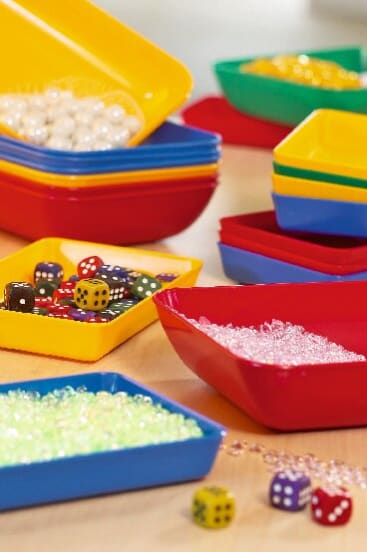 Bunte Kunststoffschalen enthalten verschiedene kleine Gegenstände, darunter mehrfarbige Würfel, weiße, hellgrüne und klare Perlen, die auf einer hellen Holzoberfläche angeordnet sind. Einige Tabletts sind im Hintergrund gestapelt.