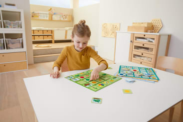 Ein junges Mädchen sitzt an einem weißen Tisch und spielt ein Brettspiel mit Bildplättchen in einem hellen, organisierten Klassenzimmer mit Holzmöbeln und Unterrichtsmaterialien im Hintergrund.