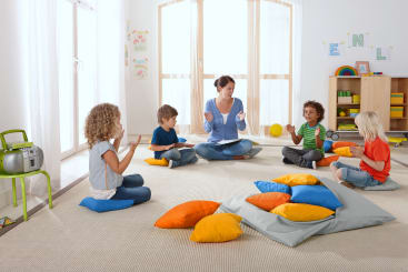 Eine Lehrerin sitzt mit vier kleinen Kindern in einem hellen Klassenzimmer auf dem Boden, umgeben von bunten Kissen. Die Gruppe scheint zu klatschen und an einer Gruppenaktivität teilzunehmen. Im Hintergrund sind Regale und Kunstwerke zu sehen.