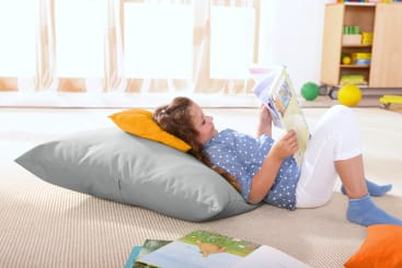 Ein Kind liegt auf einem Teppichboden, gestützt auf zwei Kissen, und liest ein Buch. Das Kind trägt ein blau gepunktetes Hemd und eine weiße Hose. In einem hellen, aufgeräumten Zimmer liegen offene Bücher und Spielzeug herum.