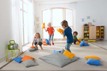 Vier kleine Kinder liefern sich eine Kissenschlacht in einem hellen, aufgeräumten Spielzimmer mit großen Fenstern. Bunte Kissen liegen auf dem Boden verstreut, und im Hintergrund stehen Regale mit Spielzeug und Büchern. Ein Radio steht auf einem kleinen Tisch.