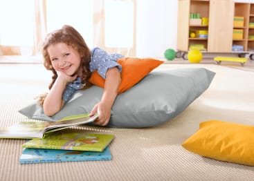 Ein junges Mädchen liegt auf einem großen grauen Kissen, stützt den Kopf auf eine Hand und lächelt, während es ein Buch liest. Mehrere bunte Bücher und Kissen sind auf dem beigen Teppich um sie herum verstreut.