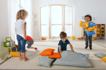Vier kleine Kinder spielen mit orangefarbenen und grauen Kissen in einem hellen Raum mit großen Fenstern. Ein Kind kniet und ordnet Kissen, ein anderes hält gestapelte Kissen, während zwei andere in der Nähe stehen und sich bücken. Im Hintergrund sind Spielzeuge und Zeichnungen zu sehen.