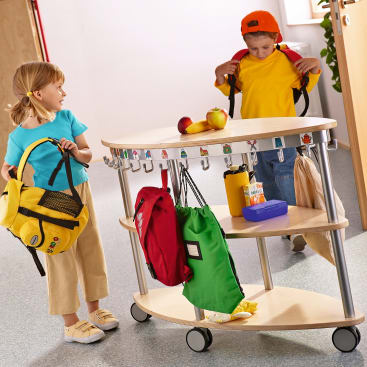Zwei Kinder stellen ihre Rucksäcke auf einen hölzernen Garderobenständer mit Haken und Fächern. Auf dem Ständer befinden sich mehrere bunte Taschen und eine Brotdose. Die Kinder scheinen sich in einem Klassenzimmer oder in einer Kinderbetreuung zu befinden.