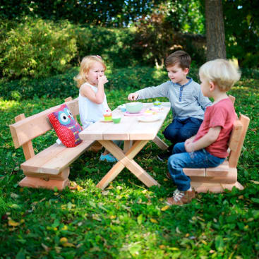 Drei kleine Kinder sitzen an einem hölzernen Picknicktisch in einem grasbewachsenen Außenbereich und essen Snacks. Ein rotes Eulenkissen liegt auf einer Bank. Die Umgebung ist von Bäumen beschattet, durch die das Sonnenlicht hindurchscheint.