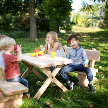 Vier kleine Kinder sitzen an einem hölzernen Picknicktisch im Freien und spielen mit Spielzeuggeschirr und Tassen an einem sonnigen Tag. Im Hintergrund sind Bäume und grünes Gras zu sehen.