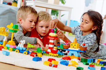 Drei kleine Kinder spielen zusammen auf dem Boden mit bunten Bauklötzen und bauen verschiedene Strukturen und Fahrzeuge auf einem weißen Teppich in einem hell erleuchteten Raum zusammen.